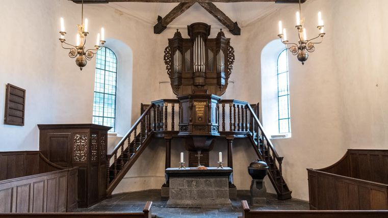 Eine einfache Innenansicht einer Kirche mit einer Orgel auf einer Empore. Es gibt Holzverkleidungen und Kerzenleuchter, die eine ruhige Atmosphäre schaffen.