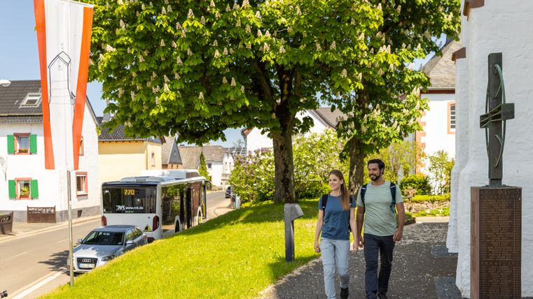 Zwei Wandernde gehen auf einem gepflasterten Weg an einer weißen Kapelle vorbei, daneben steht ein Eifelsteig-Wanderbus am Straßenrand. Im Hintergrund prägen grüne Bäume, Ortshäuser und eine Fahne die sonnige Dorfansicht.