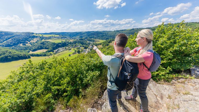Ein Paar steht auf einem Aussichtspunkt und blickt auf eine grüne Landschaft. Die Sonne scheint und der Himmel ist blau mit einigen Wolken.