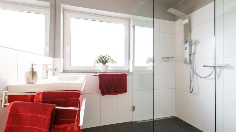 Ein modernes Badezimmer mit gläserner Dusche und hellen Fliesen. Rote Handtücher und eine kleine Pflanze verleihen dem Raum Farbe.