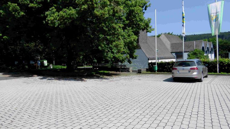 Ein Parkplatz mit gepflastertem Boden und einem Auto. Im Hintergrund sind Bäume und ein Gebäude mit Fahnen zu sehen.