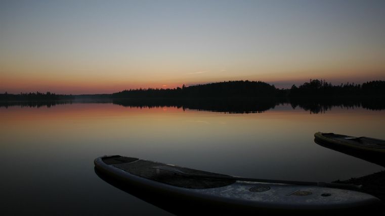 Ein ruhiger See bei Sonnenuntergang mit sanften Farben. Im Vordergrund liegen zwei Stand-Up-Paddle-Boards am Ufer.