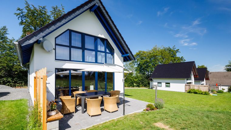 Ein modernes Haus mit großen Fenstern und einem gemütlichen Außenbereich. Der Garten ist grün und einladend, ideal zum Entspannen.