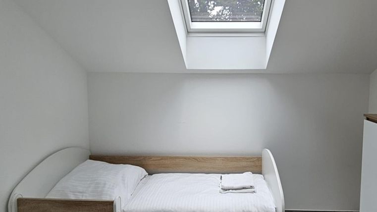 Ein einfaches Schlafzimmer mit einem Einzelbett und einem kleinen Fenster. Die Wände sind weiß und der Boden ist grau.