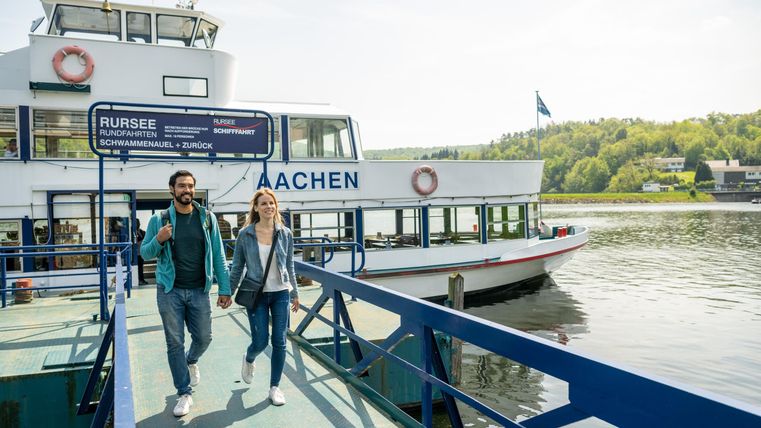 Eine Gruppe von Personen geht an einem Bootsteg entlang. Im Hintergrund ist ein Ausflugsschiff am Wasser zu sehen.