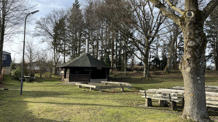 Ein ruhiger Außenbereich mit einer Laube und Sitzbänken. Der Platz ist von Bäumen umgeben und hat einen klaren Himmel.