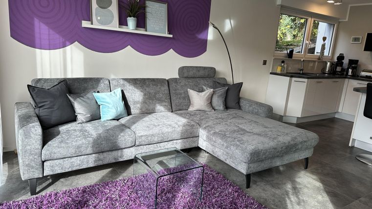 Ein modernes Wohnzimmer mit einem grauen Sofa und bunten Kissen. Der Raum hat einen lila Akzentwand und einen weichen lila Teppich.