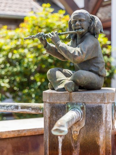 Eine bronzene Statue eines sitzenden Kindes, das Flöte spielt. Die Statue steht auf einem Brunnen mit fließendem Wasser im Hintergrund.