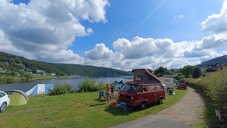 Ein schöner Campingplatz am Fluss mit einem roten Wohnmobil. Die Landschaft ist von grünen Wiesen und blauen Himmel mit fluffy Wolken umgeben.