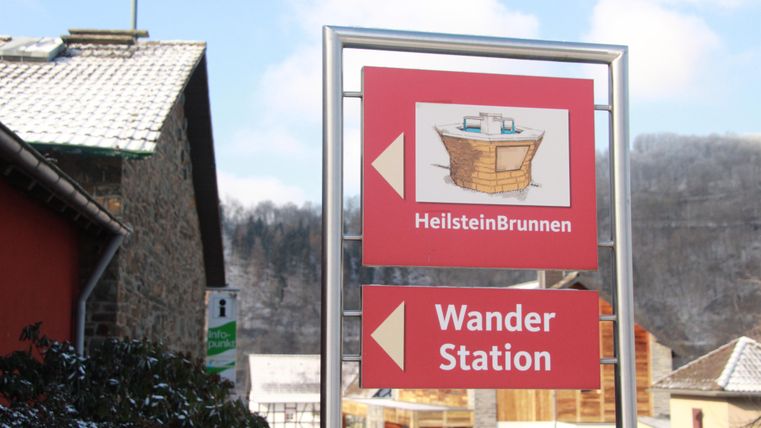 Ein Wegweiser verweist auf den HeilsteinBrunnen und die Wanderstation. Er befindet sich in einer ländlichen Umgebung mit schneebedeckten Dächern im Hintergrund.