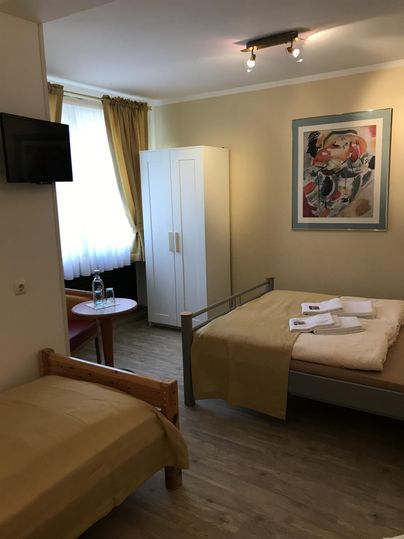 Ein einfaches Zimmer mit zwei Betten, einem Fernseher und einem Schrank. Das Zimmer hat große Fenster mit Vorhängen und ist hell eingerichtet.