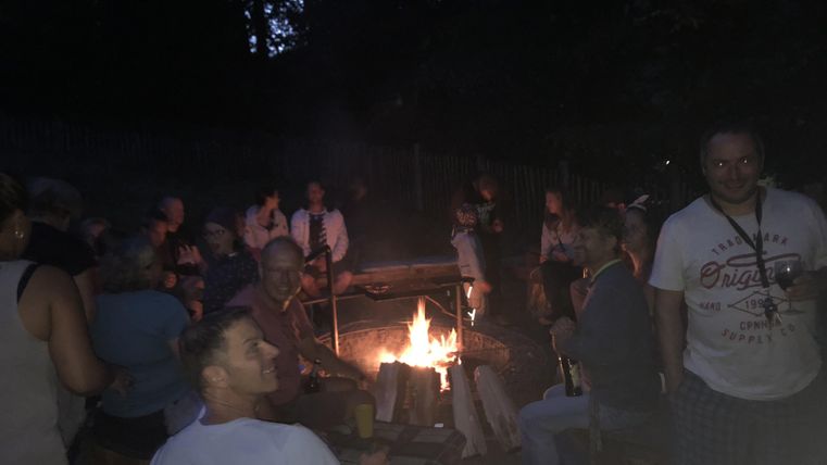 Eine Gruppe von Menschen sitzt um ein Lagerfeuer in einer dunklen Umgebung. Es herrscht eine gemütliche und gesellige Atmosphäre.