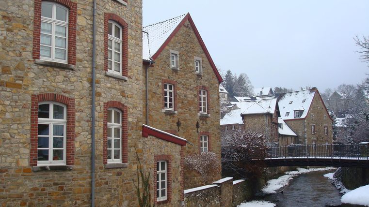 Eine idyllische, schneebedeckte Landschaft mit historischen Steinbauten. Ein ruhiger Wasserlauf verläuft entlang der Gebäude und führt zu einer kleinen Brücke.