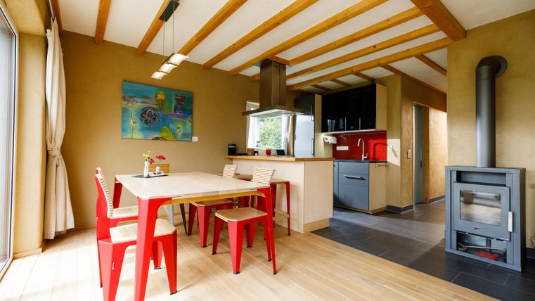 Ein modernes Esszimmer mit rotem Tisch und Stühlen. Die große Fensterfront sorgt für viel Tageslicht und eine gemütliche Atmosphäre.