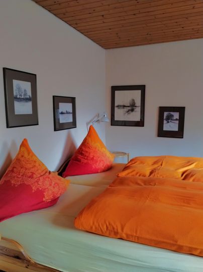Ein gemütliches Schlafzimmer mit einem großen Bett und leuchtend orangefarbenen Kissen. Die Wände sind mit verschiedenen gerahmten Bildern dekoriert.