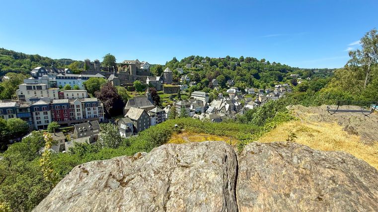 Eine malerische Aussicht auf ein kleines Dorf, umgeben von grünen Hügeln. Im Vordergrund sieht man einen großen Steinfelsen.