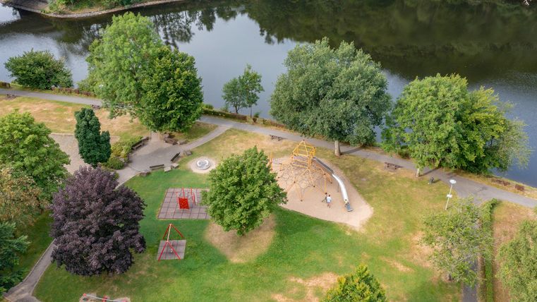 Ein grüner Park am Wasser mit vielen Bäumen. Im Park gibt es Spielgeräte und Sitzmöglichkeiten.