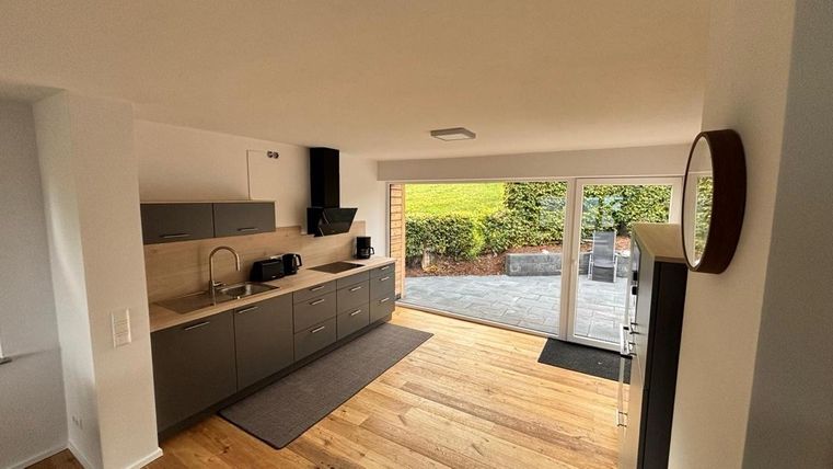 Eine moderne Küche mit grauen Schränken und Holzfußboden. Große Fenster bieten Blick auf den Außenbereich mit Garten.