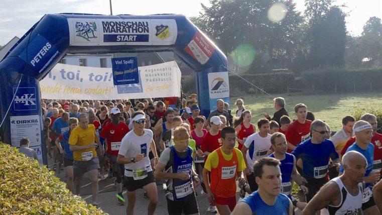 Eine große Gruppe von Läufern startet beim Monschau Marathon unter einem blauen Startbogen. Viele Teilnehmer tragen farbige Sportbekleidung und sind bereit für das Rennen.