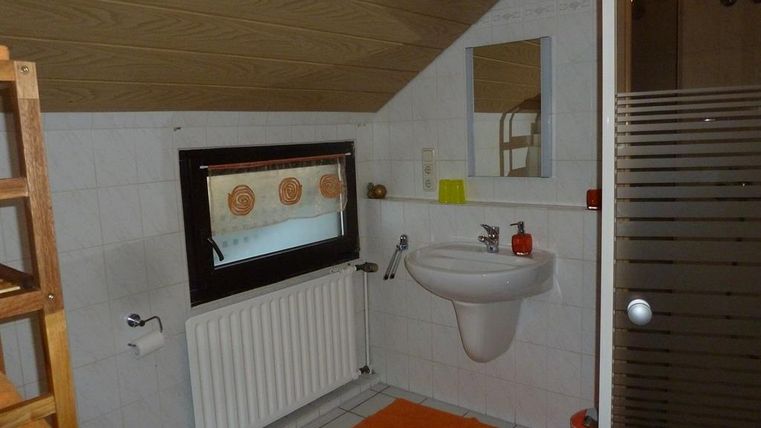 Ein kleines Badezimmer mit einem Waschbecken, einem Fenster und einer Dusche. Der Boden ist mit weißen Fliesen und einem orangefarbenen Teppich ausgestattet.