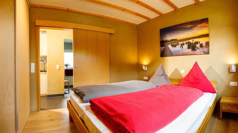 Ein gemütliches Schlafzimmer mit einem Doppelbett und roten Kissen. Die Wände sind in einem warmen Gelbton gestrichen, und ein Fenster bietet Tageslicht.