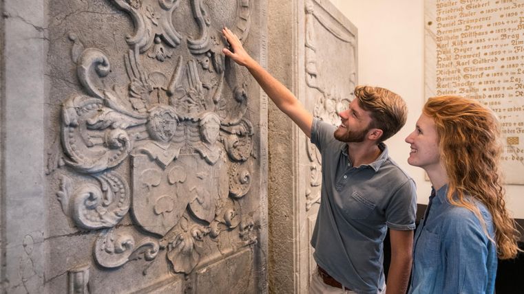 Ein Paar steht vor einer historischen Wand mit dekorativen Reliefs. Sie betrachten die Details und scheinen interessiert zu sein.