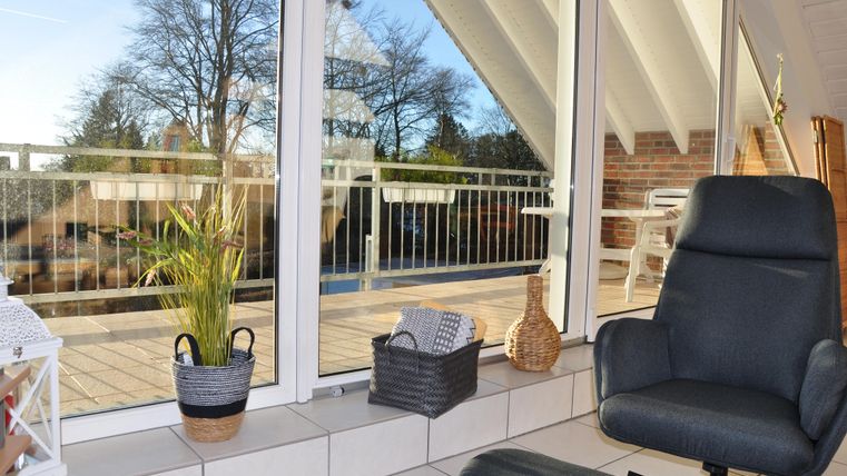 Ein gemütlicher Raum mit großen Fenstern, die auf einen Balkon mit Blick ins Freie führen. Im Vordergrund steht ein bequemer Sessel und einige Pflanzen in Körben.