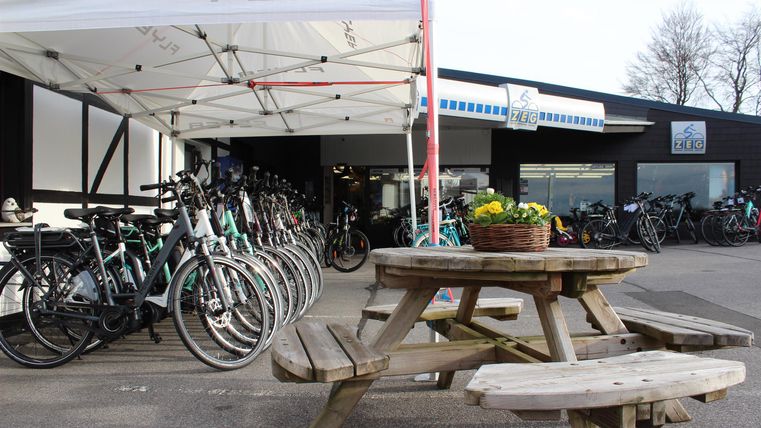 Ein Fahrradgeschäft mit einem Zelt und mehreren Fahrrädern. Im Vordergrund steht ein Holztisch mit Blumen.