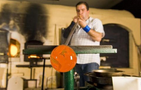 Ein Glasbläser arbeitet an einem Glasstück in einer Werkstatt. Im Hintergrund sind Öfen und Werkzeuge zu sehen.