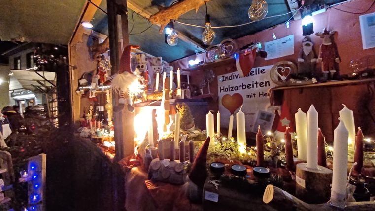 Ein festlicher Marktstand mit verschiedenen Kerzen und Dekorationsgegenständen. Die Stimmung ist warm und einladend, beleuchtet von bunten Lichtern.