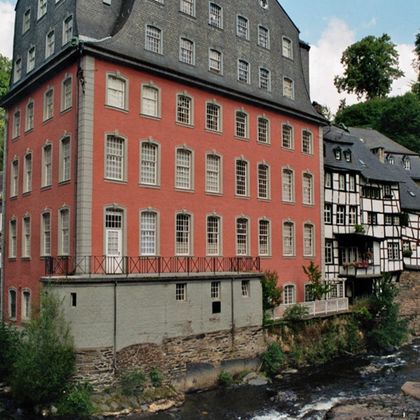 Ein rotes Gebäude neben einem Fluss in einer idyllischen Umgebung. Umgeben von traditionellen Fachwerkhäusern und grünen Bäumen.
