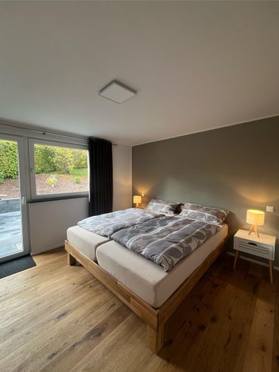 Ein gemütliches Schlafzimmer mit einem großen Bett und einem Holzboden. Es gibt Fenster mit Blick nach draußen und zwei Nachttische mit Lampen.