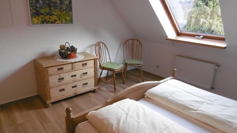 Ein einfaches Zimmer mit zwei Einzelbetten und einem Fenster, das viel Licht hereinkommen lässt. Im Raum stehen ein Holztisch und zwei Stühle sowie eine Kommode.