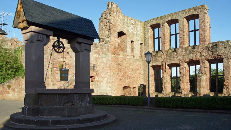 Eine alte Ruine mit großen Fenstern und einem historischen Brunnen im Vordergrund. Die Sonne scheint auf die steinernen Wände und sorgt für eine malerische Atmosphäre.