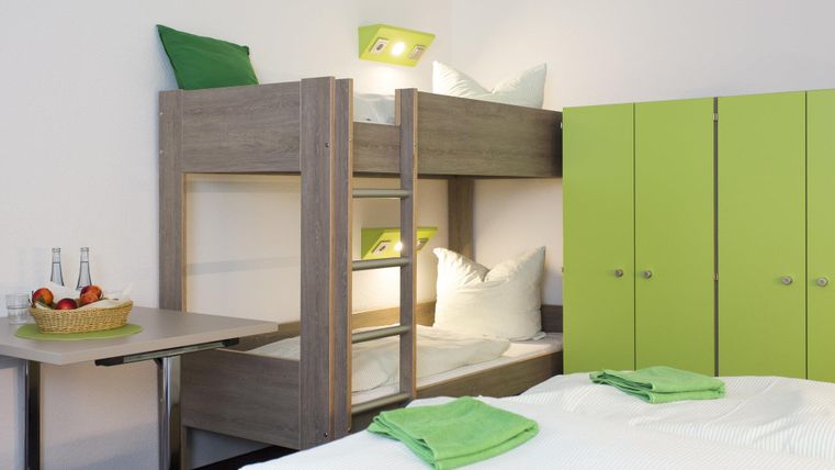 Ein funktionales Zimmer mit einem Etagenbett und einem Doppelbett. Die Möbel sind modern und in freundlichen Farben gehalten.