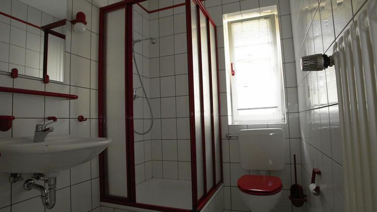 Ein kleines Badezimmer mit weißen Fliesen und roten Akzenten. Es enthält eine Dusche, ein Waschbecken und eine Toilette.