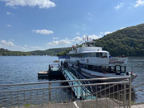 Ein Schiff liegt am Steg an einem schönen See, umgeben von grünen Hügeln. Der Himmel ist klar und blau mit einigen weißen Wolken.