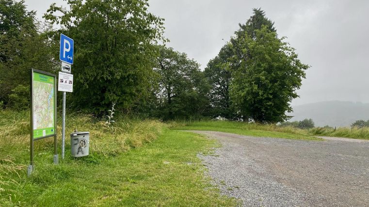 Ein Parkplatz in der Natur, umgeben von Bäumen und Gras. Der Himmel ist bewölkt, und der Weg führt ins Grüne.