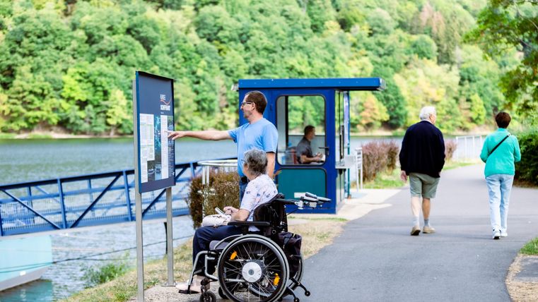 Eine Person im Rollstuhl schaut auf eine Informationstafel am Ufer eines Gewässers. Im Hintergrund gehen zwei weitere Personen auf einem Spazierweg.