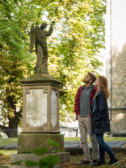 Ein Paar steht vor einer Statue eines Engels in einem Park. Umgeben von Bäumen in leuchtenden Farben genießen sie den Anblick.