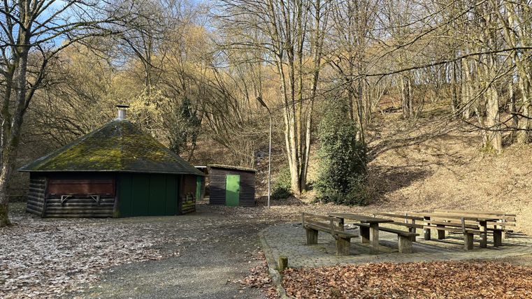 Eine Hütte in einem Waldgebiet mit Laubbäumen. Neben der Hütte steht ein Grillplatz mit Sitzbänken.