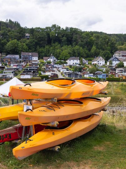 Drei stapelbare, orangefarbene Kajaks stehen auf einer Wiese. Im Hintergrund sind kleine Häuser und Bäume zu sehen.
