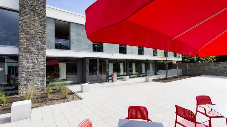 Ein moderner Innenhof mit roten Sonnenschirmen und Tischen. Die angrenzende Fassade des Gebäudes ist mit großen Fenstern gestaltet.