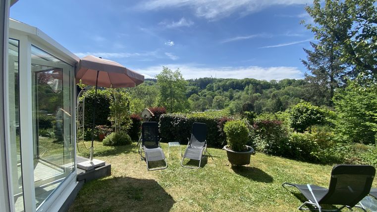 Ein schöner Garten mit Liegestühlen und einem Sonnenschirm. Im Hintergrund sind Hügel und Bäume unter einem klaren blauen Himmel sichtbar.