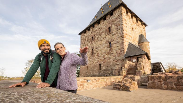 Ein Paar steht lächelnd vor einem historischen Turm. Sie zeigen auf die Architektur und genießen den Moment.