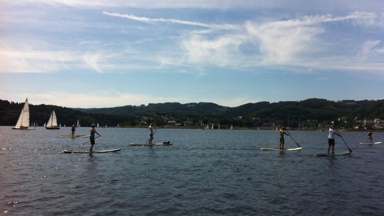 Eine Gruppe von Menschen steht auf Stand-Up-Paddling-Boards auf einem ruhigen See. Im Hintergrund sind Segelboote und sanfte Hügel zu sehen.