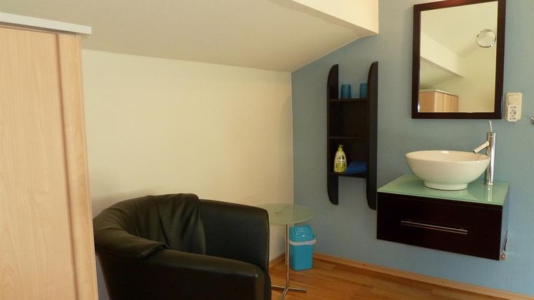 Ein kleiner Raum mit einem schwarzen Sessel und einem Waschbecken. Die Wände sind blau gestrichen und es gibt einen Spiegel sowie ein Regal.