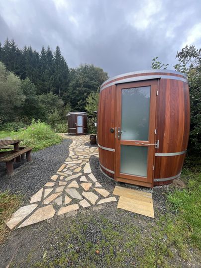 Zwei holzverkleidete Saunafässer stehen in einer naturnahen Umgebung. Ein gepflasterter Weg führt zu den Fässern, umgeben von grünen Bäumen und Sträuchern.