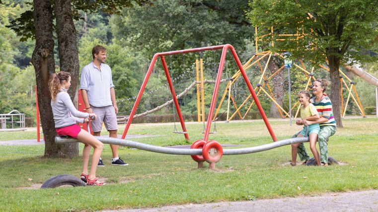Ein Spielplatz mit einer Wippe und zwei Gruppen von Kindern. Im Hintergrund sind Schaukeln und viel grünes Gras zu sehen.