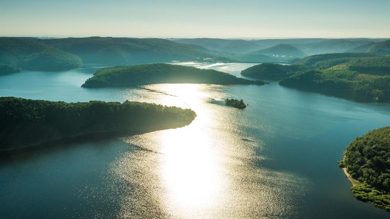 Ein malerischer See mit sanften Hügeln und üppigem Grün. Das Sonnenlicht spiegelt sich glänzend auf der Wasseroberfläche.